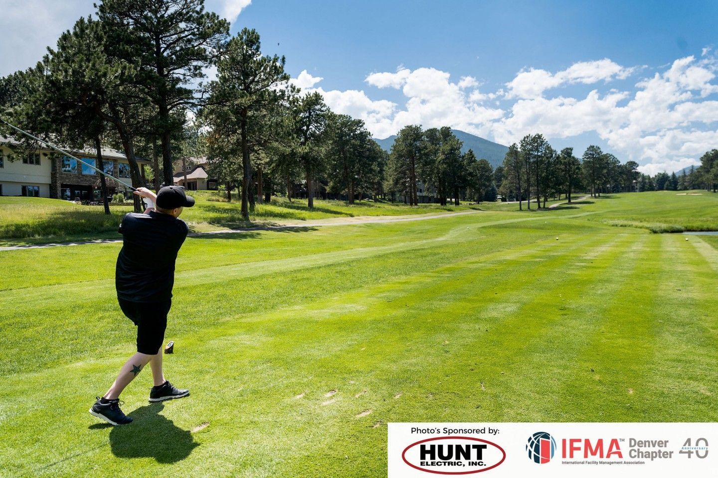 Golfer swinging on a sunny golf course, trees and mountains in the background.
