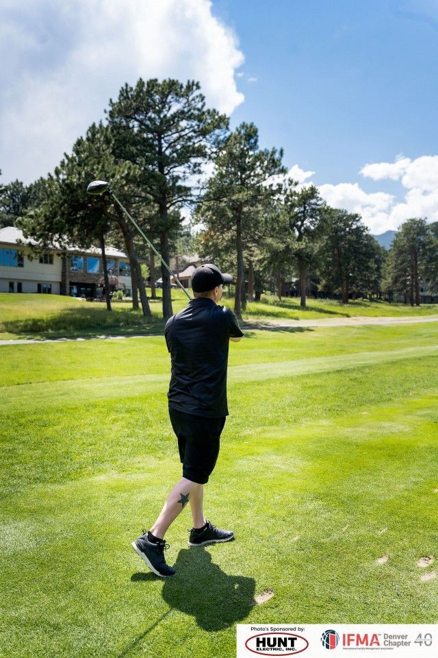 Golfer swinging a club on a sunny green golf course. Trees and buildings are visible in the background.