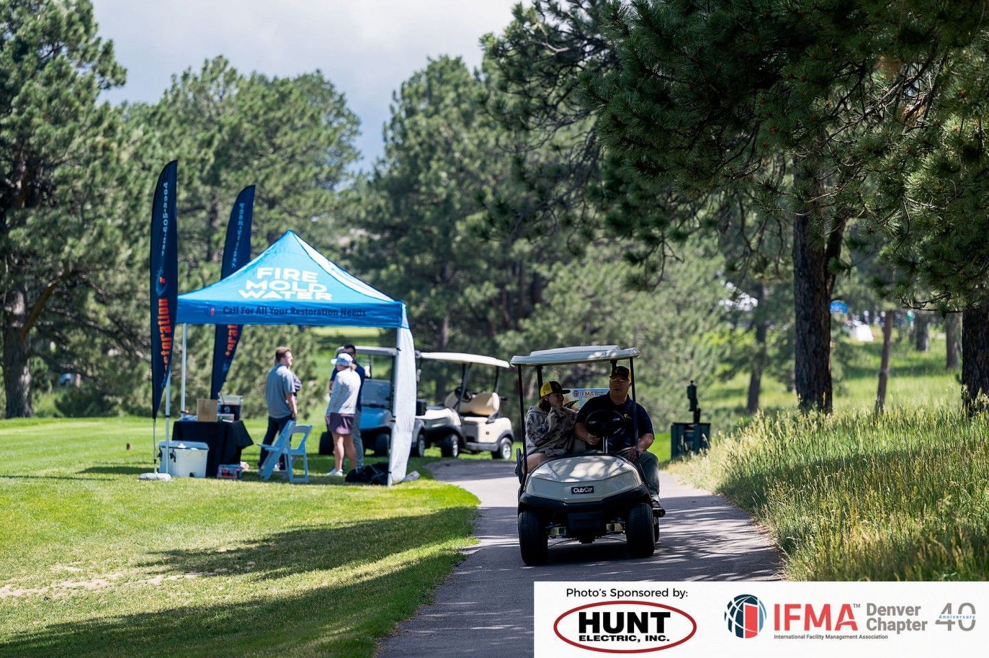 Golfers in golf cart on pathway near tent during an event.