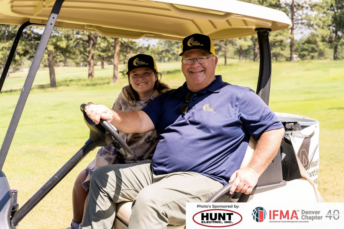 Man and person in a golf cart on a course; both wearing hats and smiling.