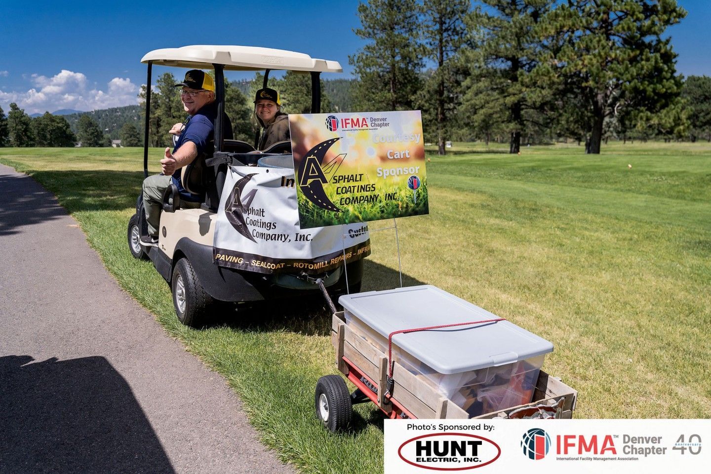 Golf cart with two people towing a trailer carrying a container, driving on a golf course.