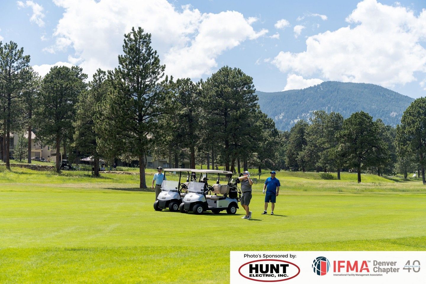 Golfers on a green course with golf carts, trees, and a mountain backdrop under a partly cloudy sky.