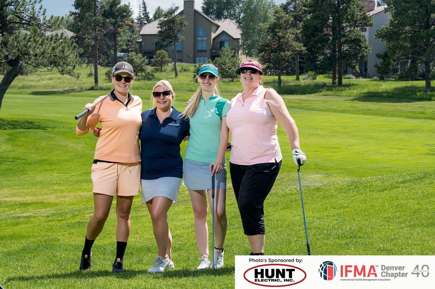 Four women pose on a golf course, smiling, holding clubs, wearing golf attire.