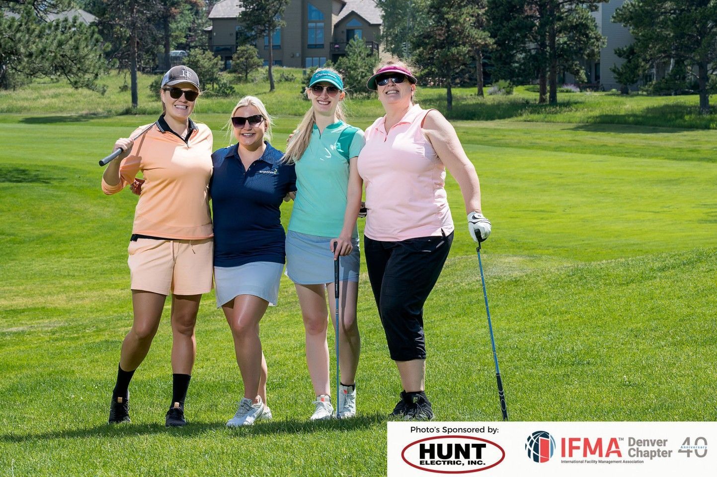Four women on a golf course, smiling, holding clubs, wearing golf attire.