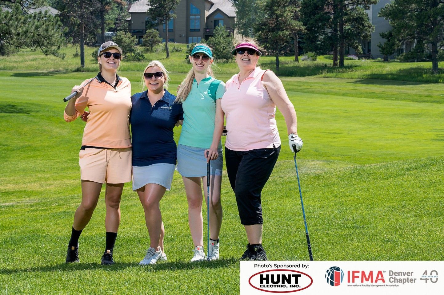Four women on a golf course, smiling, holding clubs, wearing golf attire, sunny day.