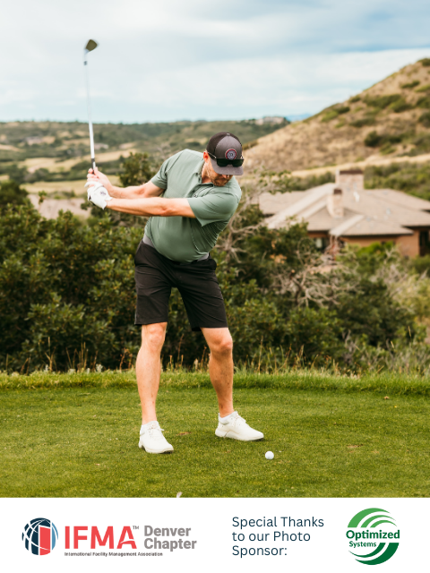 Man swinging a golf club on a green, ball on the ground. Mountains and a house are in the background.