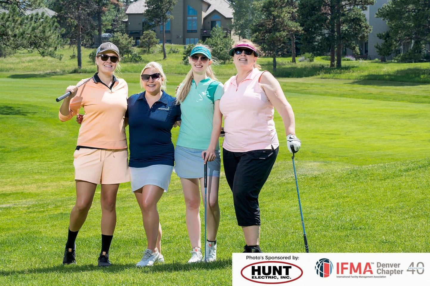 Four people on a golf course smiling, holding clubs, wearing golf attire.