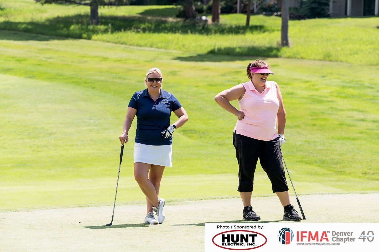Two women on a golf course, smiling, holding clubs. One in a blue shirt and white skirt, the other in pink.