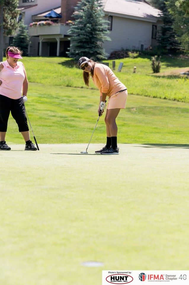 Two women on a golf course: One watches another put a golf ball. Green grass and sunny day.