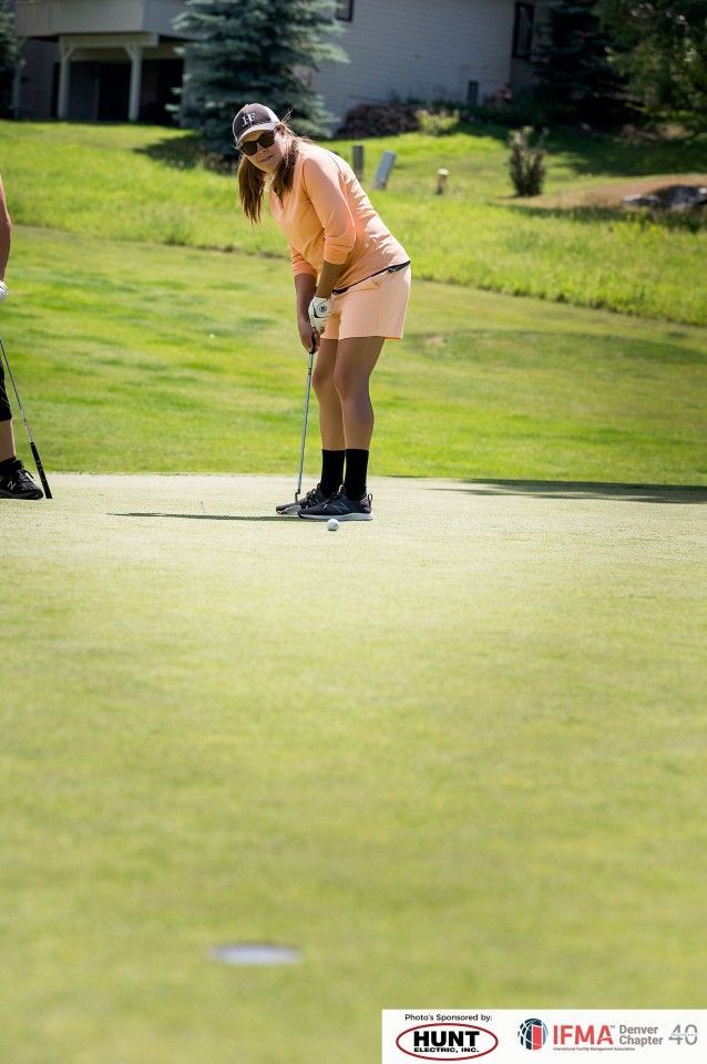 Woman golfing, putting on a green lawn. She wears an orange outfit, sunglasses, and a hat. Sunny day.