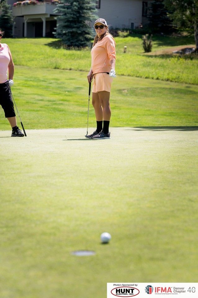 Woman in peach golf attire, putting on a green. Another golfer waits nearby.