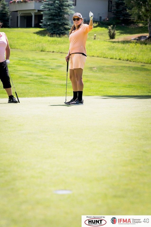 Woman on a golf course raising her arm, holding a golf club, wearing an orange outfit.