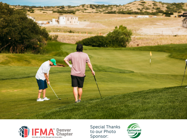 Two golfers on a green course, one putting, in front of a landscape with hills and buildings.