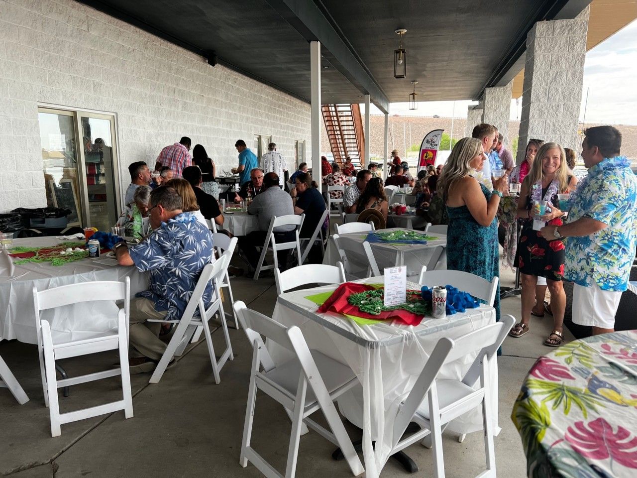 Outdoor party with people at tables. Some wear Hawaiian shirts. Under a covered patio.