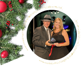 Man and woman holding an award in a circular frame; decorated with Christmas greenery and red ornaments.