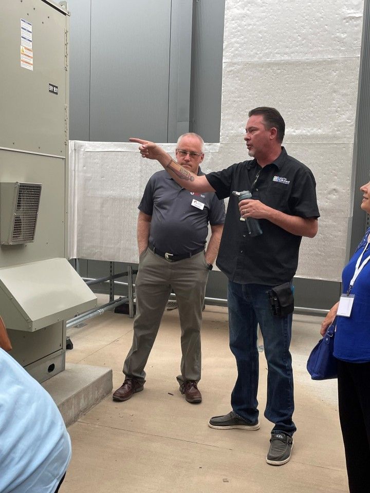 Man pointing at HVAC unit, explaining to a small group in a mechanical room.