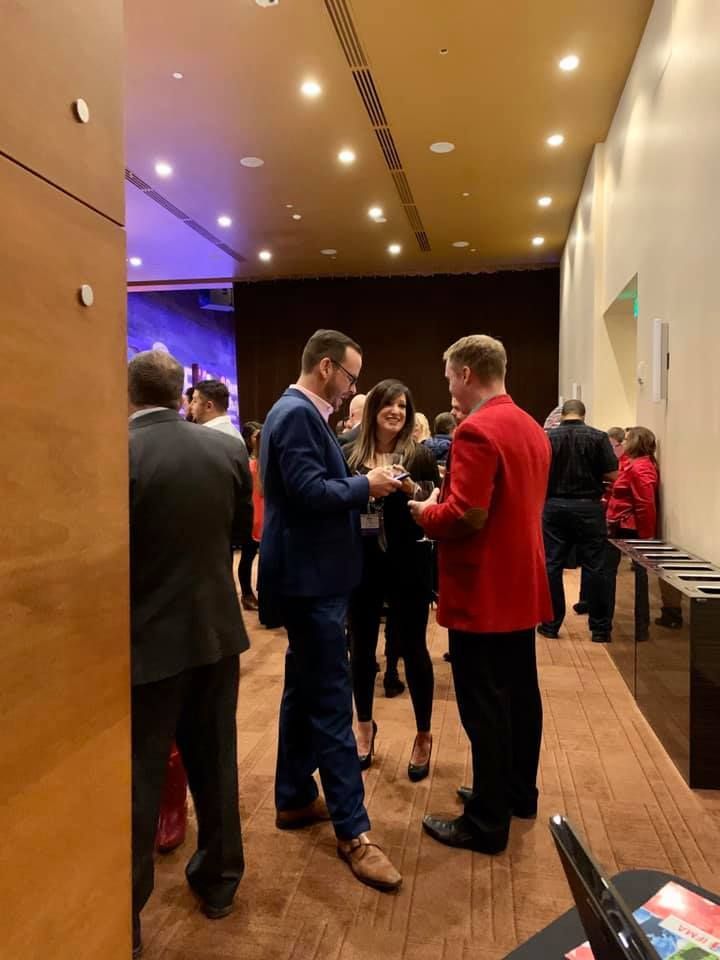 People in business attire chatting in a well-lit event space. One person in a red jacket.