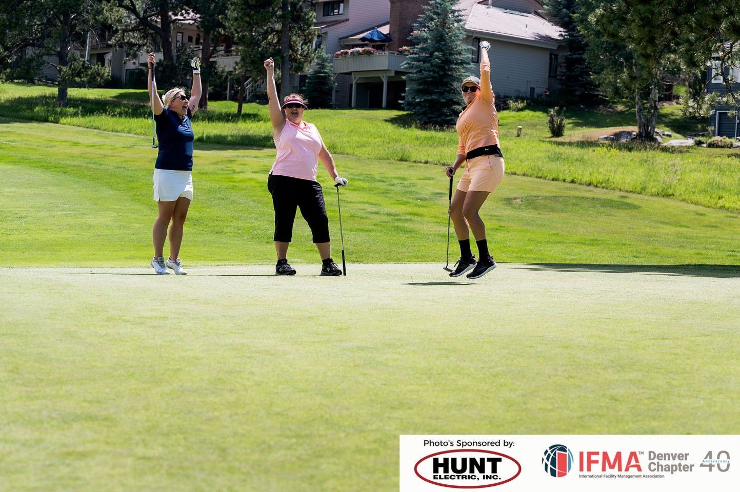 Three women on a golf course celebrating, arms raised. Green grass, sunny setting.