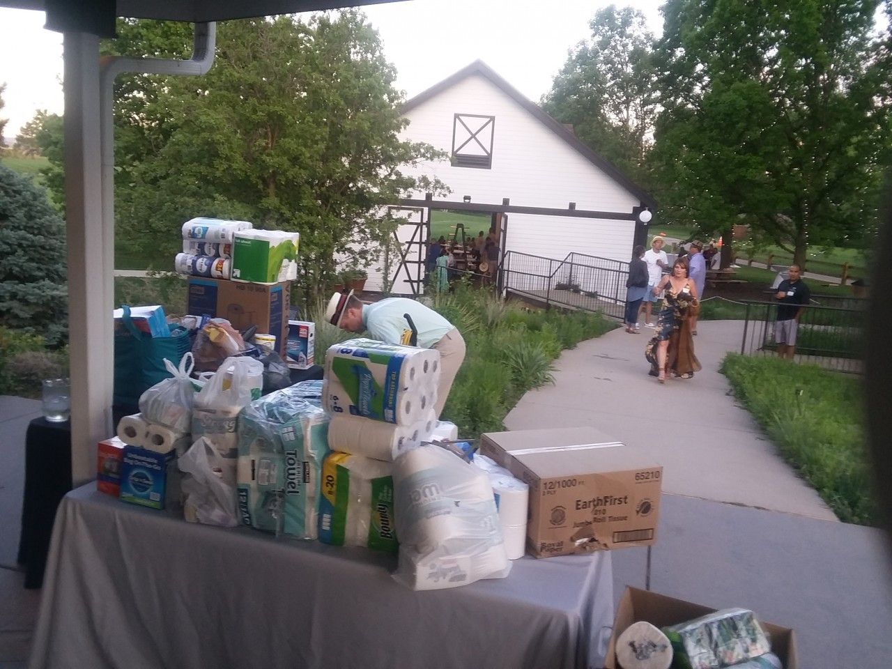 Table with supplies like paper towels, diapers. Woman sorting supplies near a white barn, people on a path.