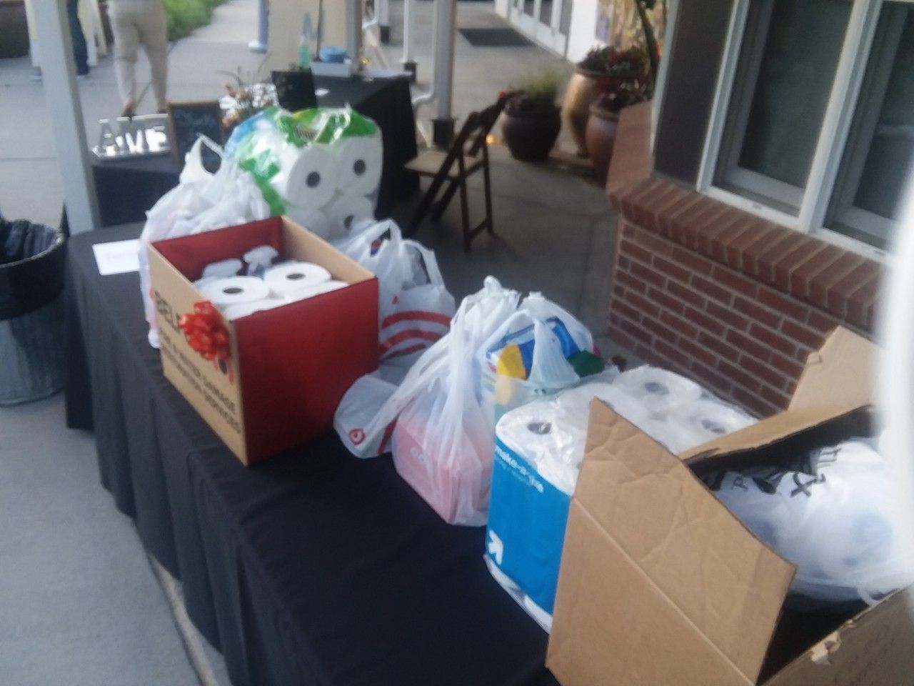 Table with donated supplies: toilet paper, paper towels, and other items. Outdoors, near a building.