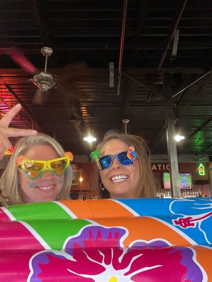 Two women smiling with sunglasses above colorful surfboards. One gives a peace sign.