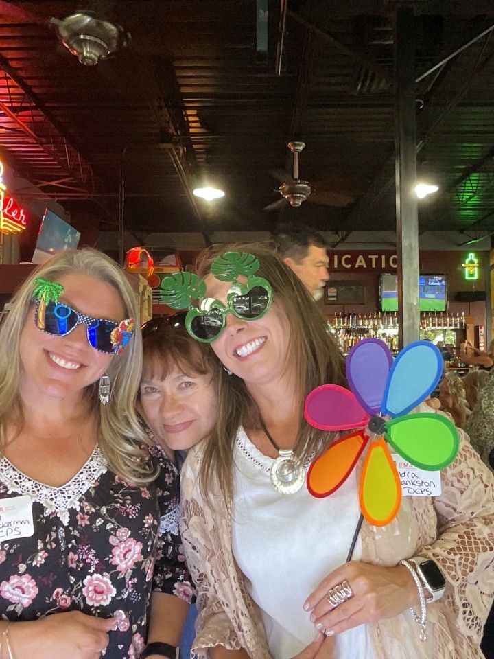 Three women smiling, wearing novelty sunglasses and holding a pinwheel at an event.