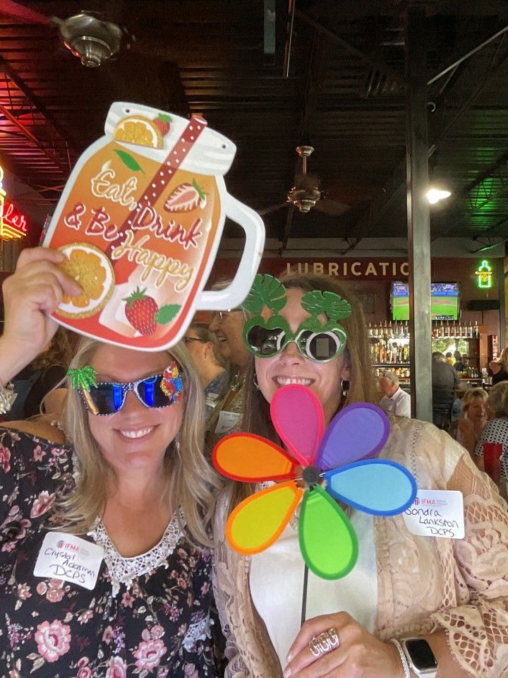 Two women smiling, holding photo props. One holds a drink sign, the other a colorful pinwheel and glasses.