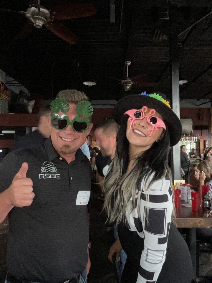 Two people smiling with novelty glasses at an indoor event, man giving thumbs up.
