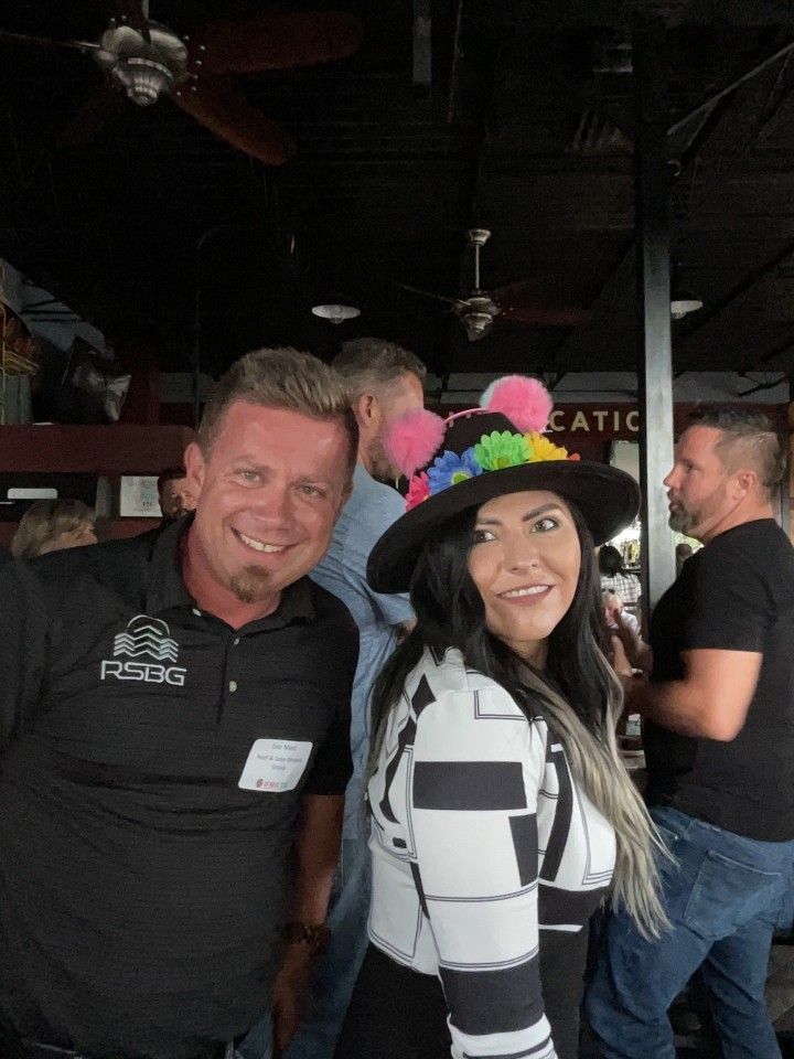 Man and woman smiling at an outdoor gathering. Woman wears a decorative hat.