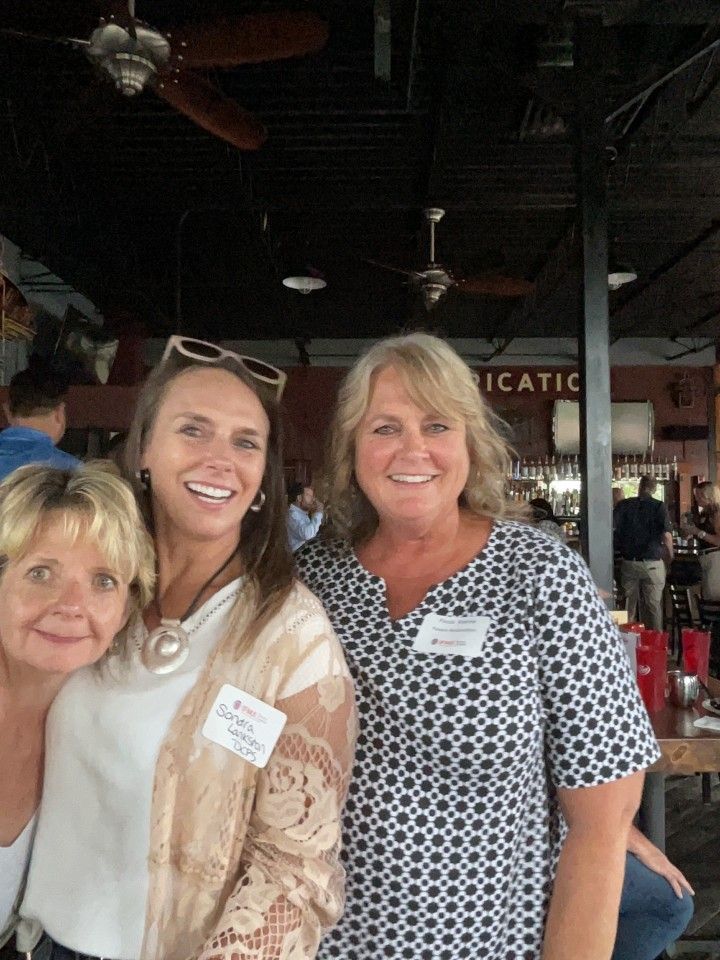 Three smiling women at a gathering, near a bar. One wears a patterned dress, another has a light sweater.