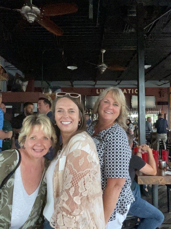 Three smiling women pose outdoors at a restaurant. One wears a lace top; the others, printed blouses.