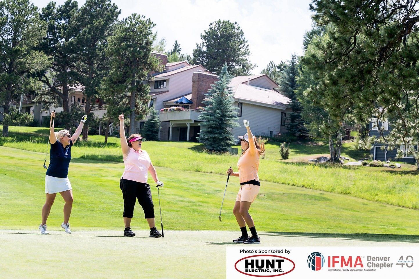 Three women celebrate on a golf course, raising their arms. Green grass, trees, and buildings in the background.