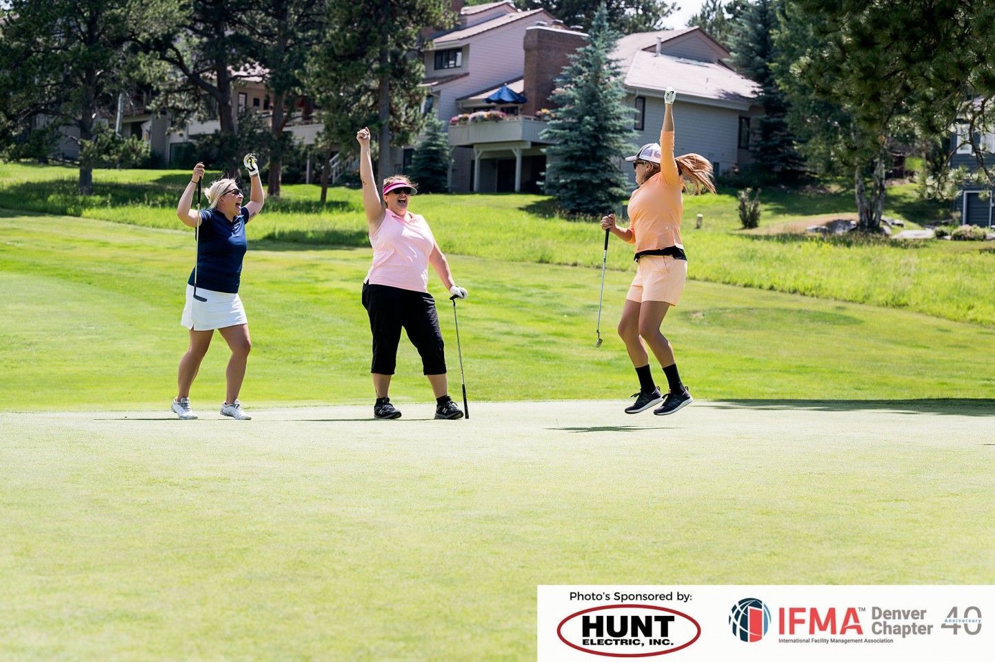 Three women celebrating on a golf course. One jumps, all raise arms. Green grass, blue sky.