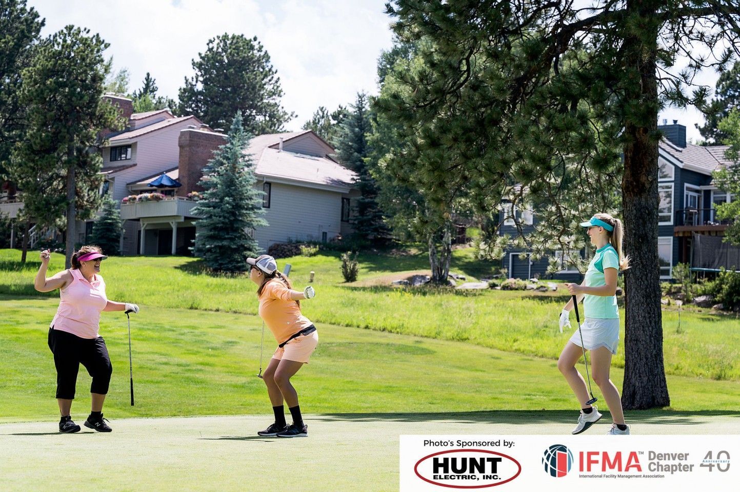 Three people playing golf on a green; one raises arms in celebration, others swing clubs. Buildings in background.
