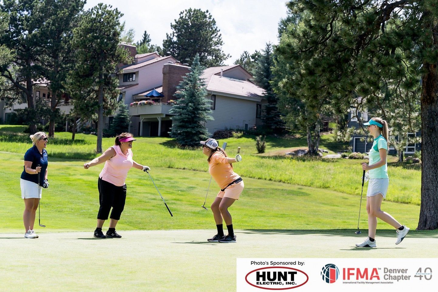 Women on a golf course; one laughing as another prepares to swing. Green grass and trees in a sunny setting.