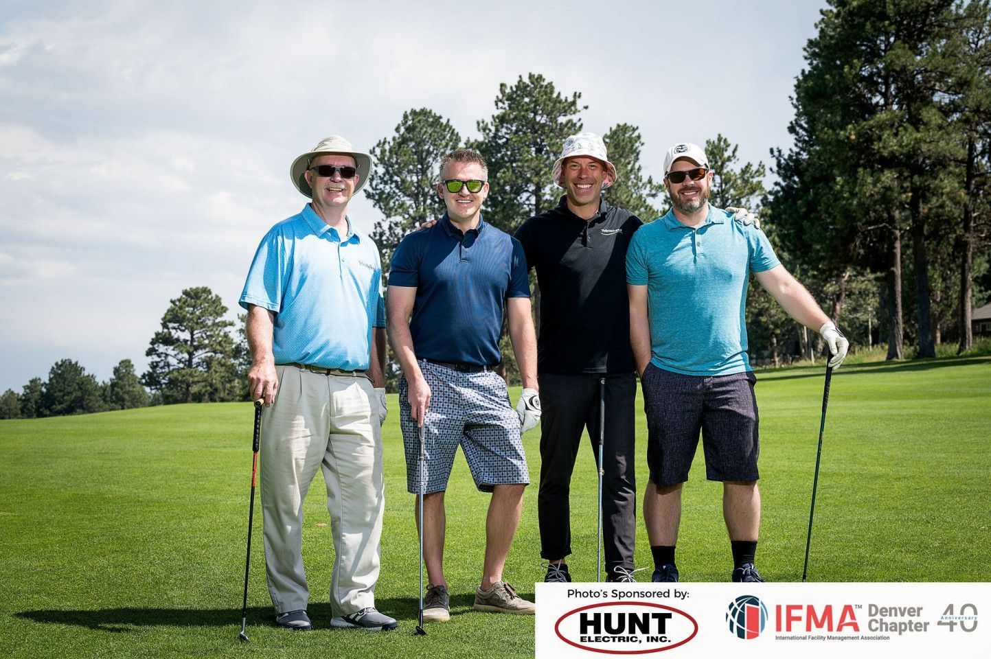 Four men on a golf course pose with clubs. Sunny day, green grass, light blue sky.