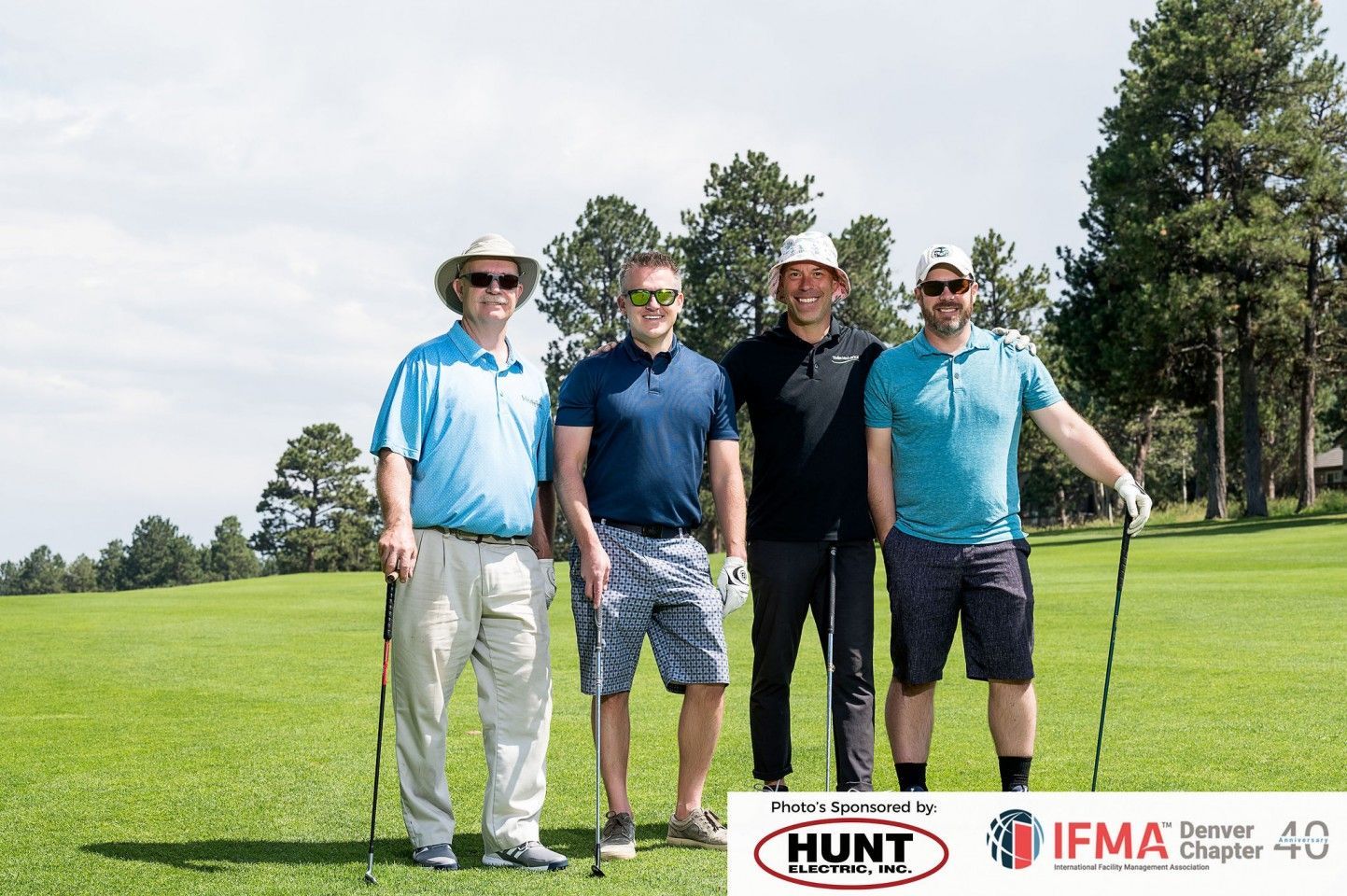 Four men on a golf course, smiling and holding clubs. Sunny day, green grass, and trees.
