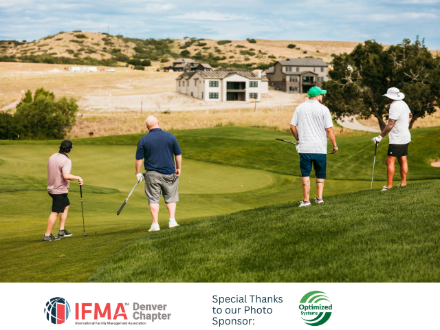 Golfers on a green course, preparing to putt, with houses and hills in the background under a cloudy sky.