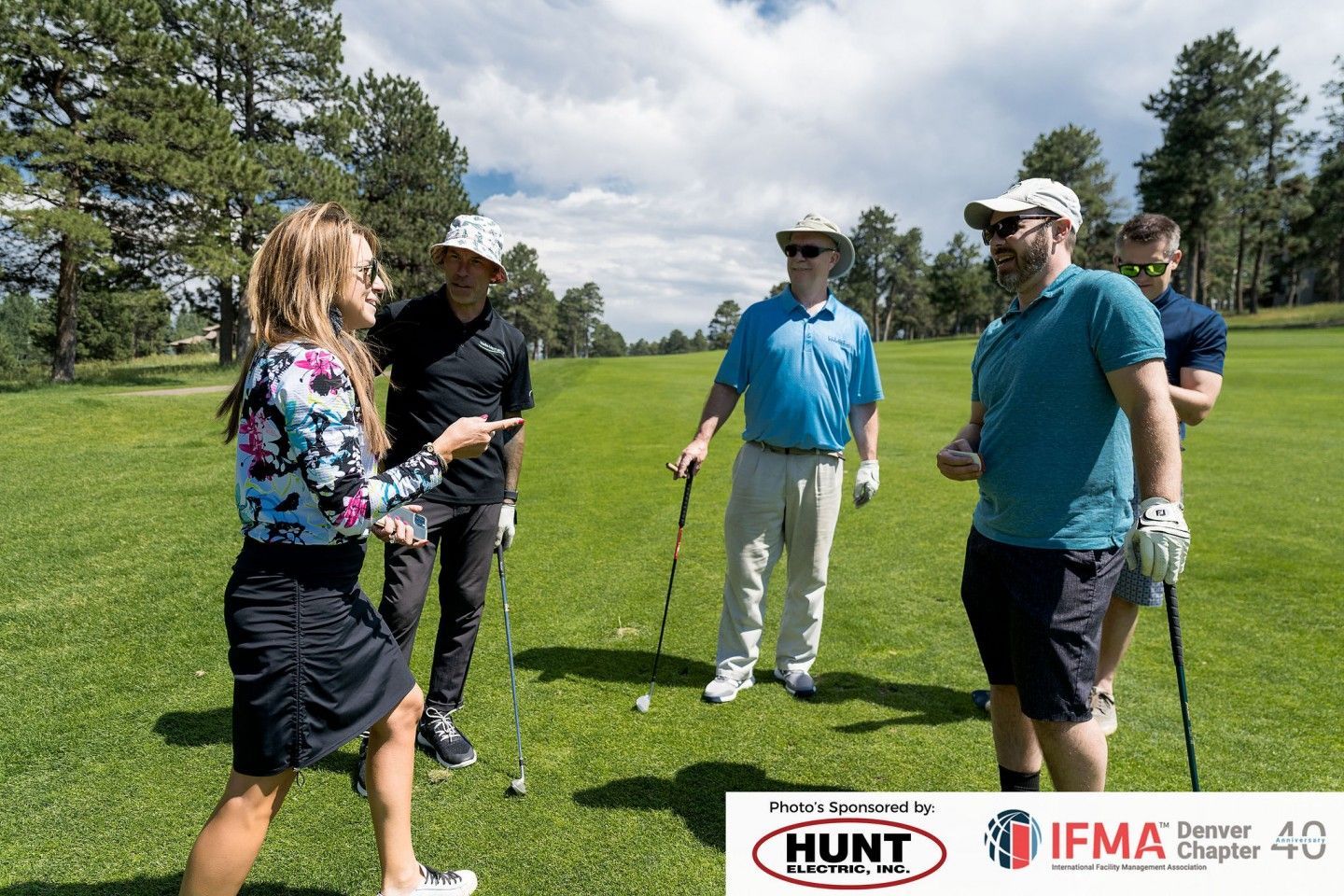 People on a golf course. A woman in a floral shirt speaks to men holding golf clubs. Sunny day.