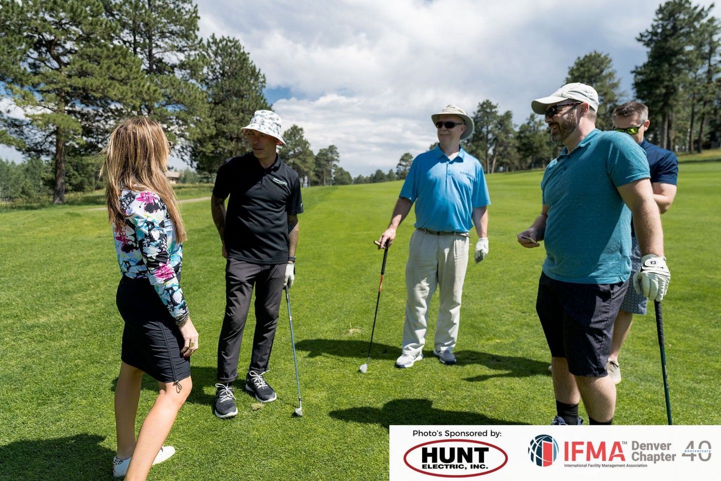 People on a golf course. Several are in a group, and one person is speaking to them. Sunny day, green grass.