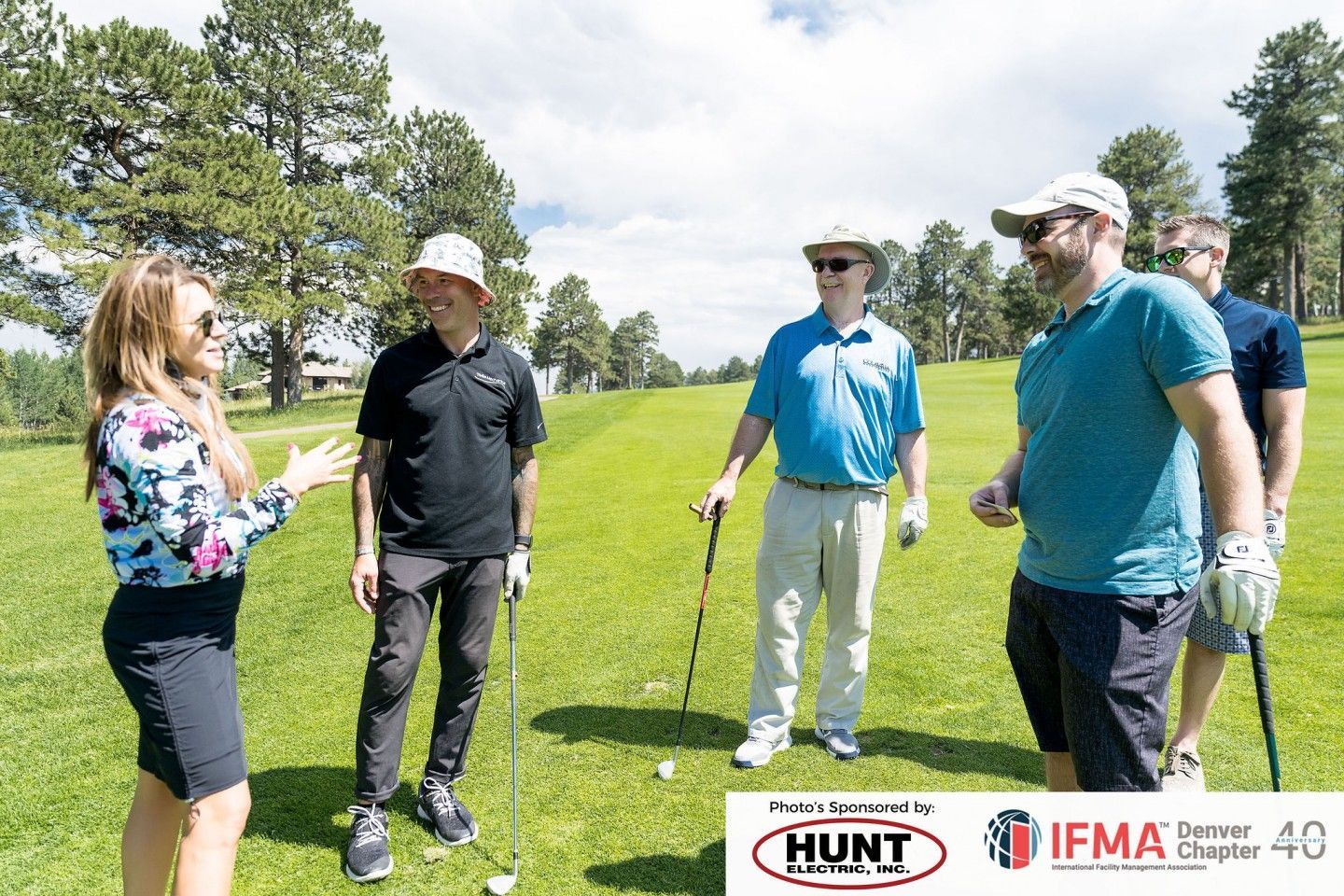 Woman speaking to a group of men on a golf course. They stand on green grass under a sunny sky.