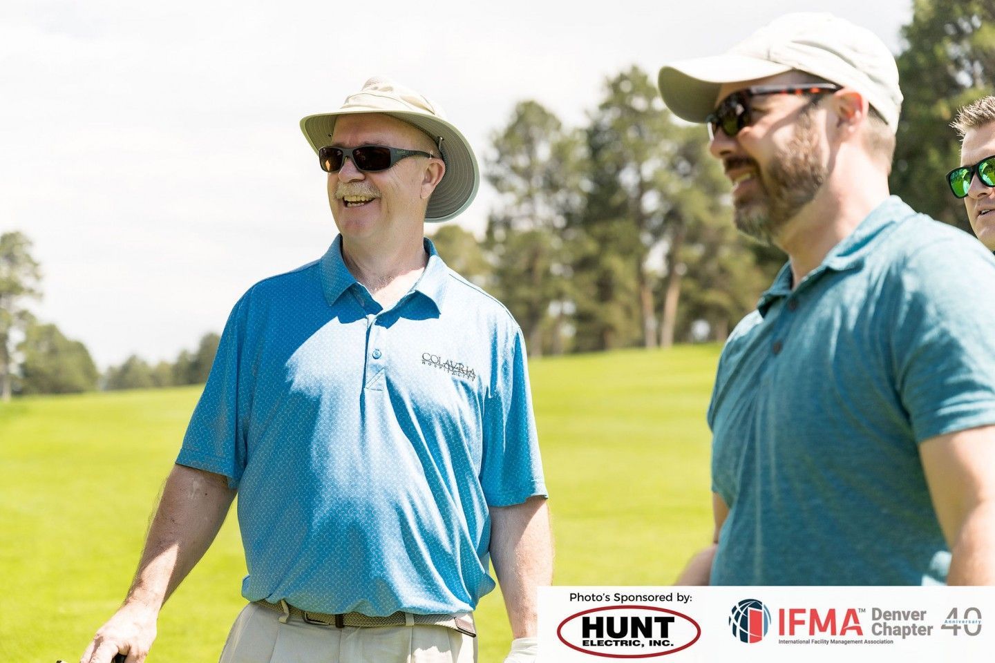 Men on a golf course wearing sunglasses and blue shirts, smiling in the sunlight.