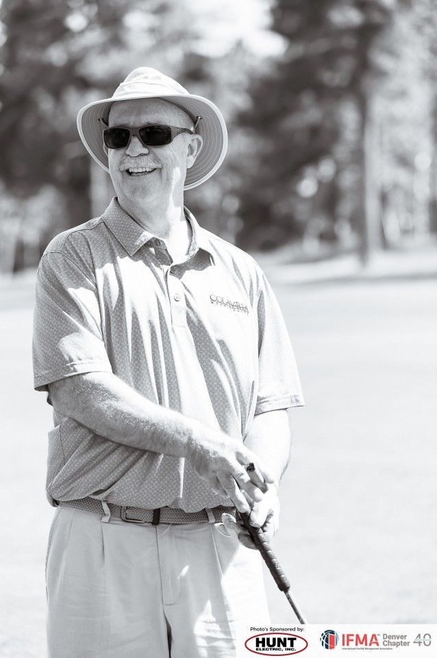 Man in sunglasses and hat, smiles while holding a golf club on a sunny golf course.