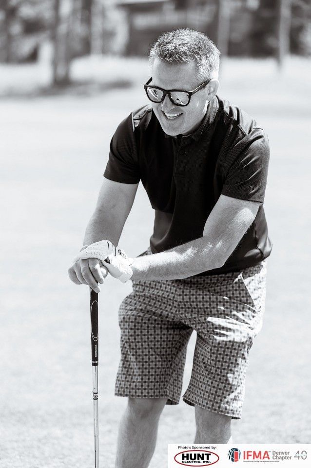 Man in eyeglasses smiles, holding golf club on a green, wearing a dark polo and patterned shorts.
