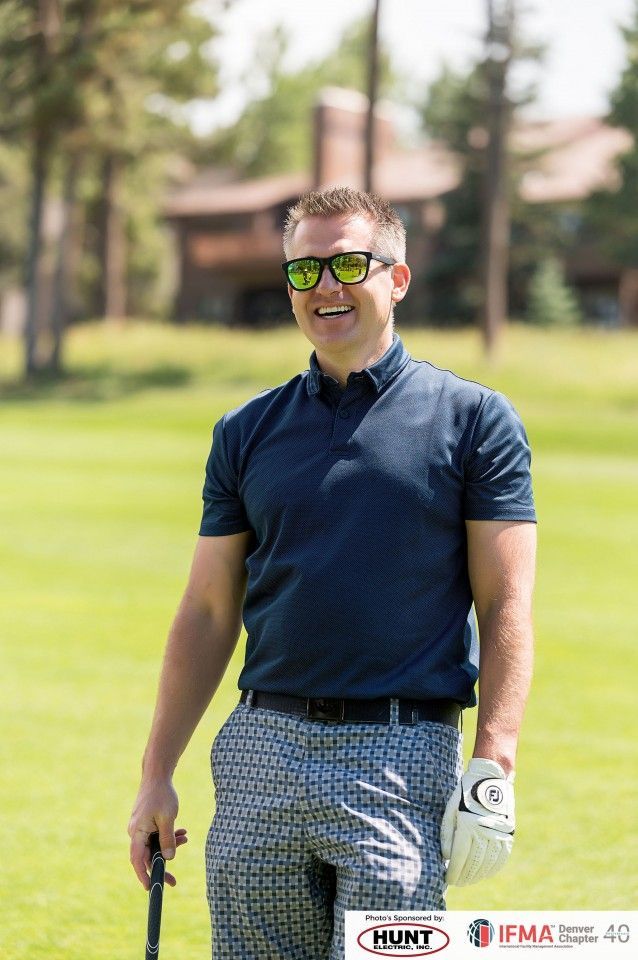 Man on golf course wearing sunglasses, navy shirt, patterned pants, holding a golf club, and smiling.