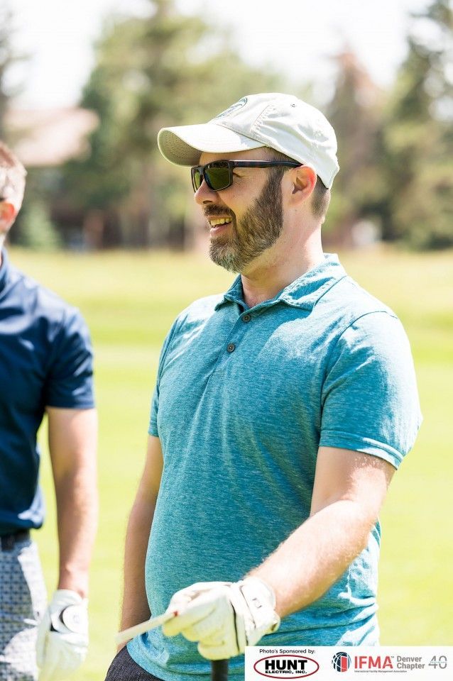 Man in blue polo shirt and cap smiles, holding a golf club on a sunny green field.