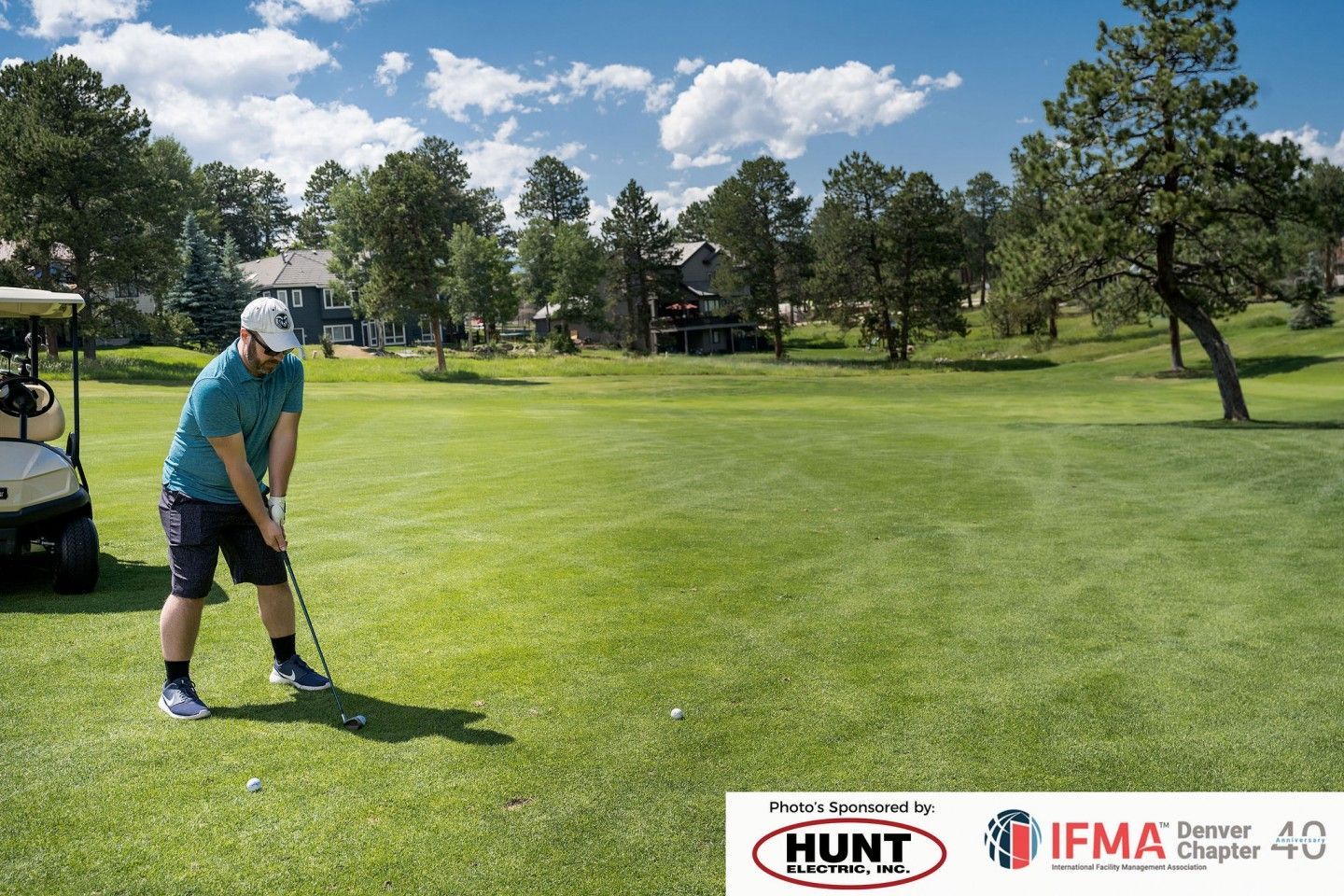 Man swings golf club on a green course, cart nearby, blue sky.