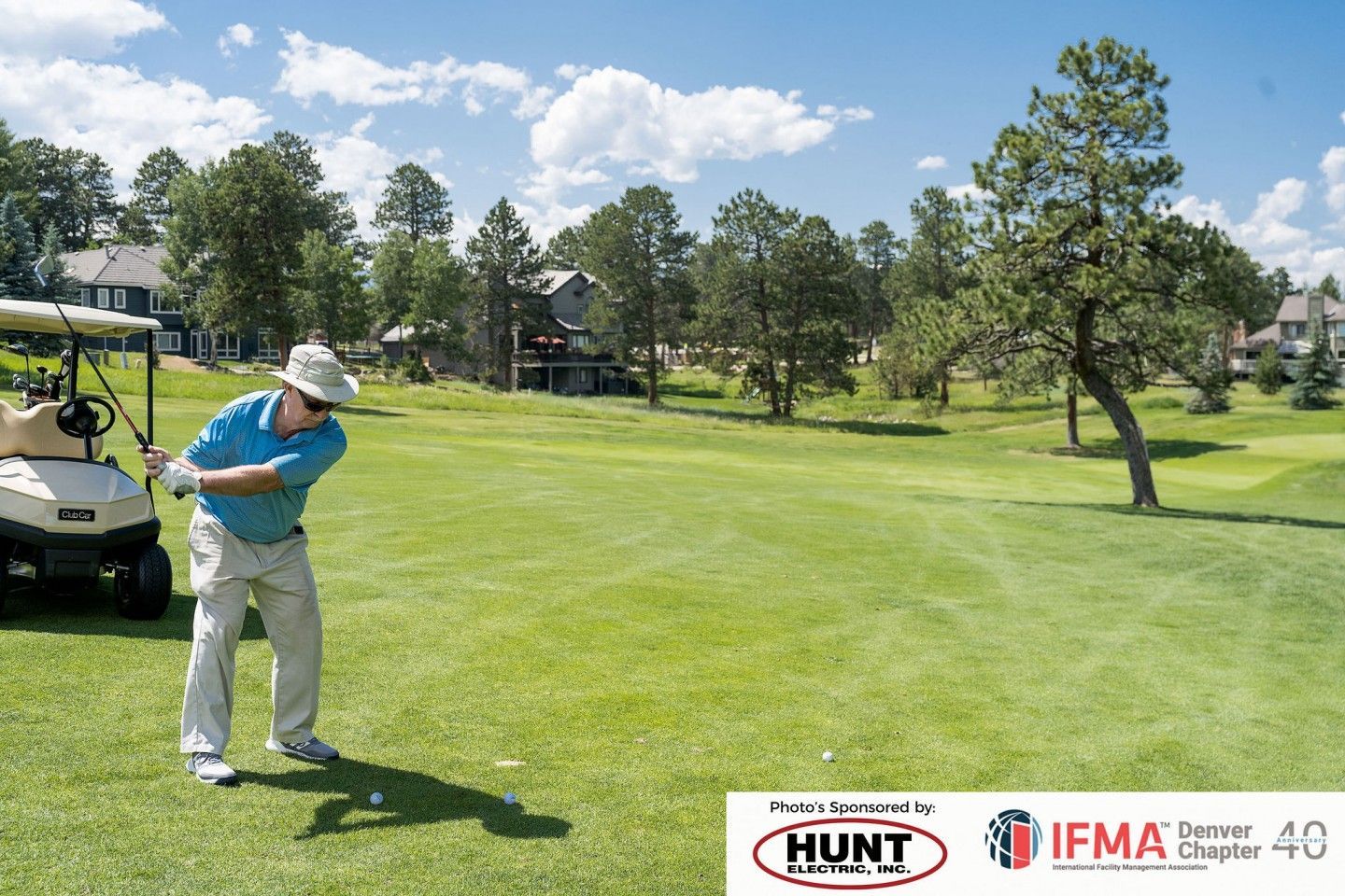 Man swinging a golf club on a green course. A golf cart and houses are in the background under a blue sky.