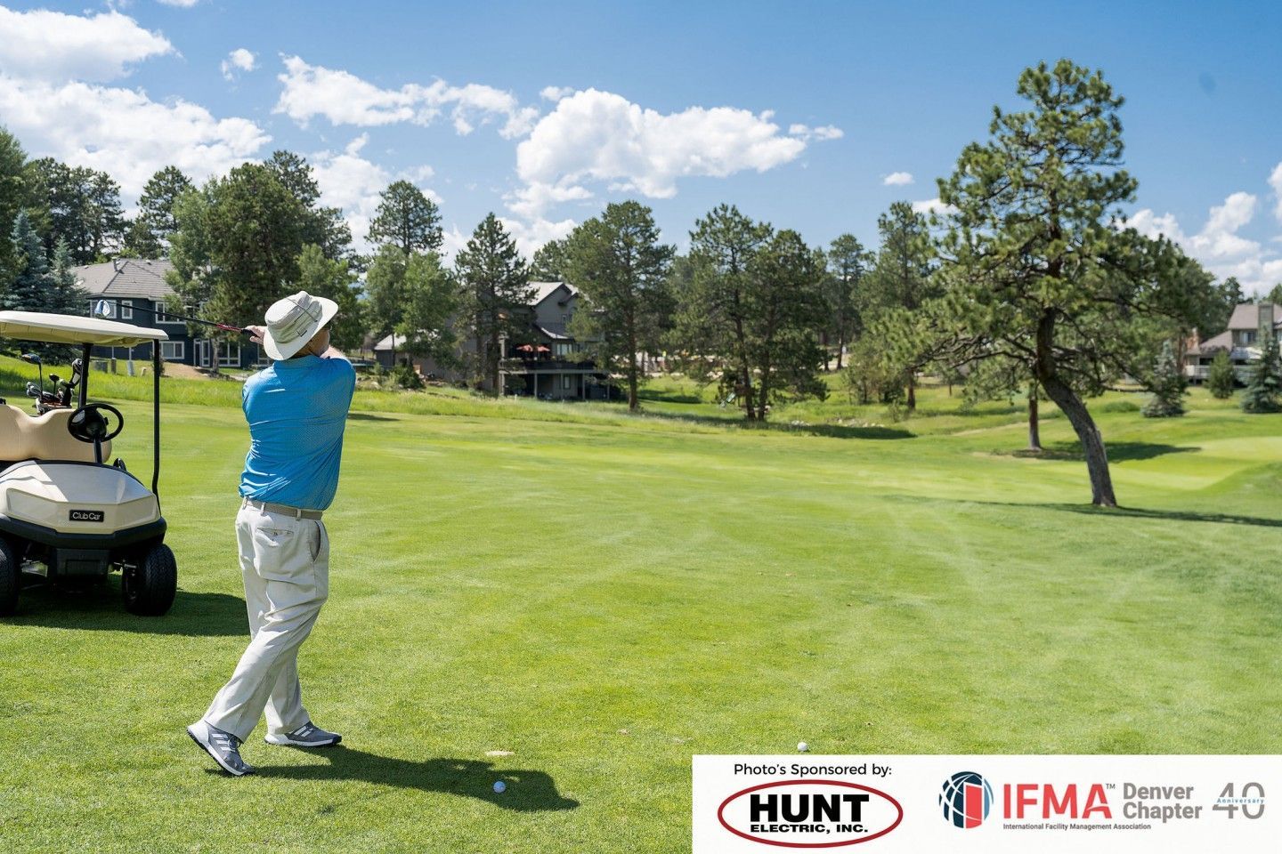 Golfer in blue shirt swings club on green course with cart and houses under blue sky.