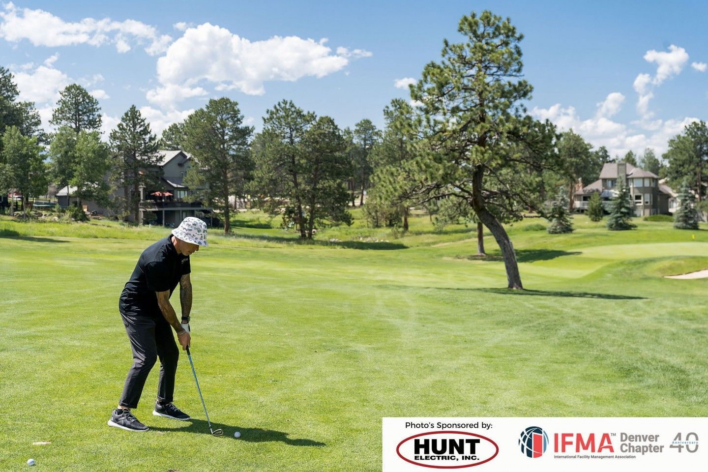 Man in black attire swings a golf club on a green course with trees and houses under a blue sky.